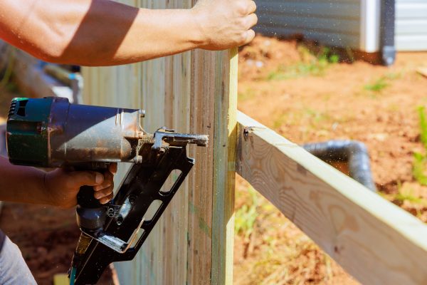 Using an air nail gun man constructs portions of wooden fence around his yard